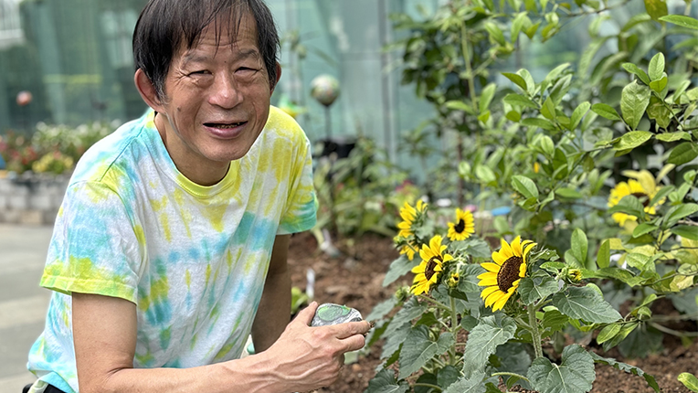 老闆潮文穿著扎染襯衫,手中握著一朵向日葵,展現出陽光般的笑容。